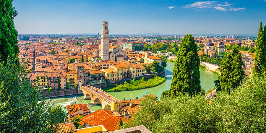 The historic city of Verona consists of red tile roofed buildings and the Ponte Pietra bridge that stretches across the green Adige river. 