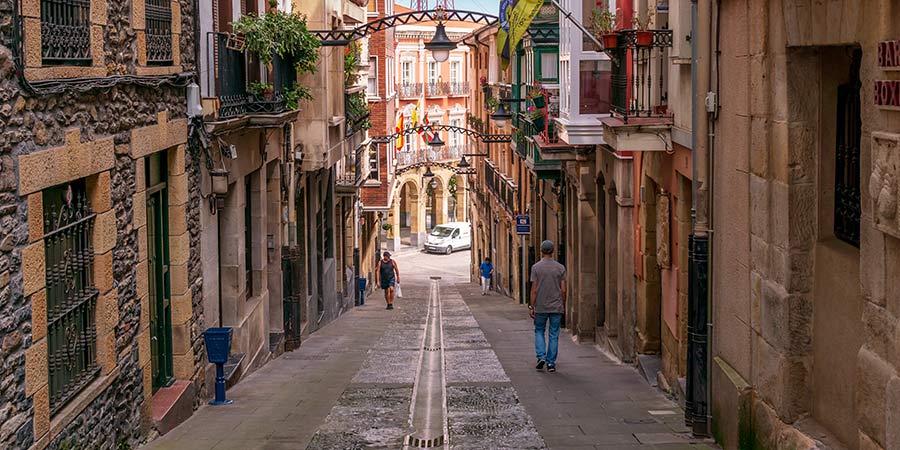 A narrow street in Bibao, traditional stone adorns the buildings and the red suspension bridge rises over the horizon.  